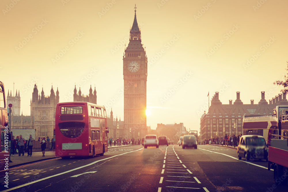 Czerwony piętrowy autobus na Westminster Bridge w Londynie, z widokiem na Big Ben i Pałac Westminsterski o zachodzie słońca. Ciepłe światło tworzy nostalgiczny klimat, a na ulicy widać ruch samochodów i pieszych.