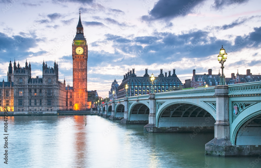 Big Ben i Pałac Westminsterski w Londynie o zmierzchu, widziane z perspektywy Tamizy. Po prawej stronie znajduje się most Westminster Bridge, którego latarnie są już oświetlone. Woda w rzece odbija subtelne światła budynków i nieba.