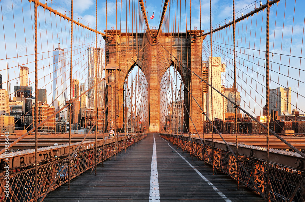 Brooklyn Bridge w Nowym Jorku o zachodzie słońca, widziany z perspektywy pieszej kładki. W tle widoczne są wieżowce Dolnego Manhattanu, w tym One World Trade Center, otoczone ciepłym światłem zachodzącego słońca.