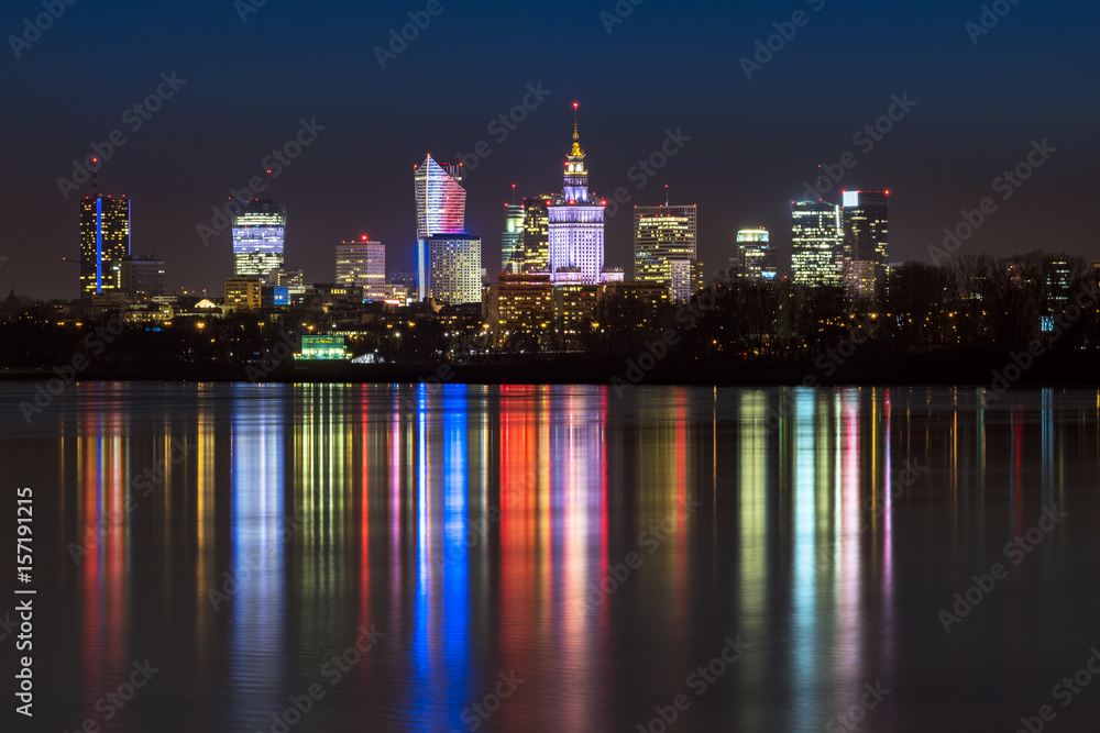 Nocna panorama Warszawy odbijająca się w gładkiej tafli Wisły - wielobarwne światła drapaczy chmur tworzą spektakl kolorowych refleksów na wodzie, z dominującym Pałacem Kultury w centrum kompozycji