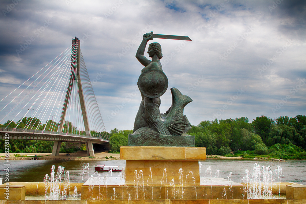 Kultowa Syrenka Warszawska na tle mostu Świętokrzyskiego - monumentalna rzeźba syreny z mieczem i tarczą, otoczona fontannami, na pierwszym planie nowoczesny most wantowy i zielone nabrzeże Wisły