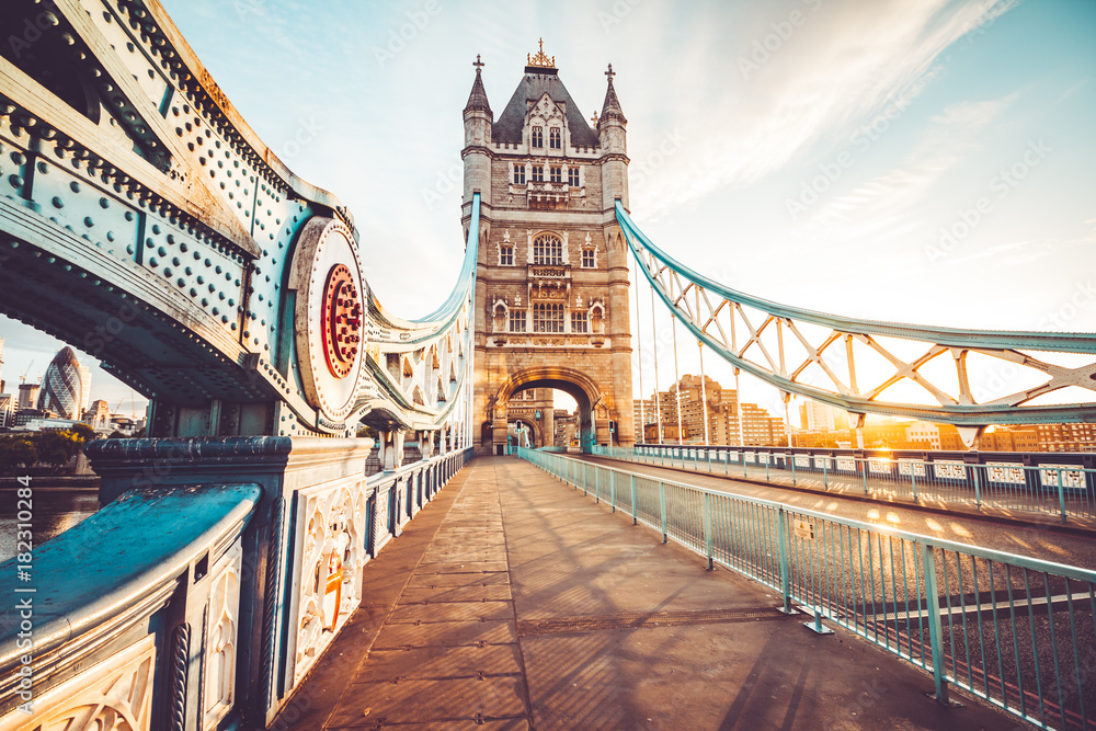 Tower Bridge w Londynie o wschodzie słońca, widziany z perspektywy pieszej kładki. Detale architektoniczne mostu, takie jak stalowe elementy i wieże, są podkreślone ciepłym światłem poranka.