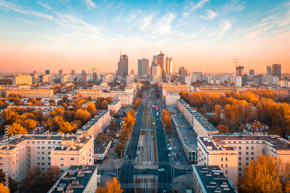 Panorama Warszawy o wschodzie słońca, długa aleja Jerozolimskie prowadzi wprost do centrum biznesowego, jesienne drzewa w złotych barwach otulają socrealistyczną zabudowę, różowo-błękitne niebo dodaje scenie ciepła i optymizmu