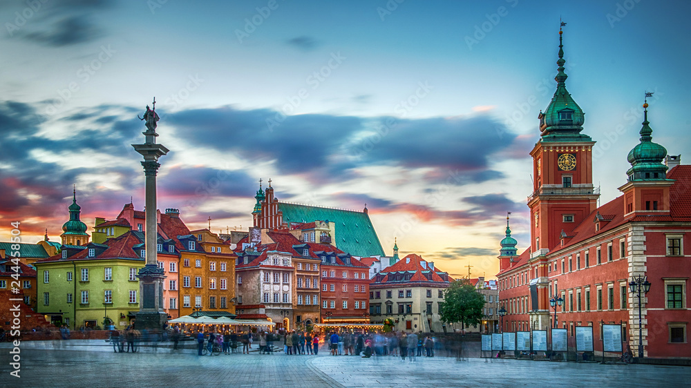 Plac Zamkowy w Warszawie o zachodzie słońca, historyczne kamienice w intensywnych kolorach, Kolumna Zygmunta i Zamek Królewski na tle dramatycznego nieba w odcieniach różu i błękitu, z tłumem ludzi tworzącym dynamiczną atmosferę miejsca