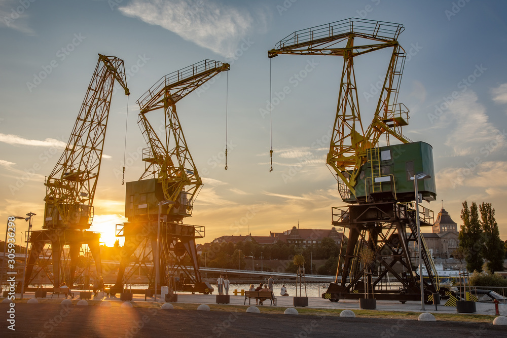 Zabytkowe dźwigi portowe w Szczecinie o zachodzie słońca - monumentalne konstrukcje w złotej poświacie, tworzące industrialną poezję na tle płonącego nieba, z charakterystyczną sylwetką miasta w tle