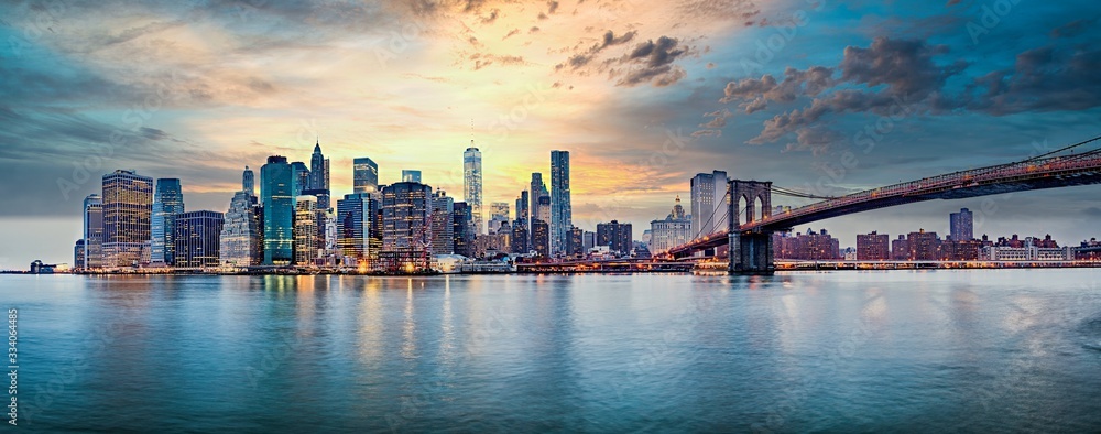 Panorama Dolnego Manhattanu w Nowym Jorku o zachodzie słońca, z widokiem na Brooklyn Bridge po prawej stronie. Wieżowce odbijają ciepłe barwy nieba w spokojnych wodach East River, tworząc harmonijną kompozycję.