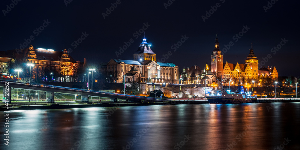 Nocna panorama Szczecina z odbiciami w wodzie - monumentalne gmachy Wałów Chrobrego rozświetlone ciepłym światłem, tworzące świetlną symfonię na spokojnej tafli rzeki, z futurystycznym mostem na pierwszym planie