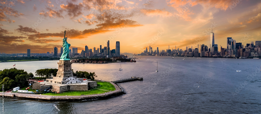 Statua Wolności na pierwszym planie z panoramą Nowego Jorku o zachodzie słońca. W tle widoczne są wieżowce Dolnego Manhattanu, w tym One World Trade Center, oraz spokojne wody rzeki Hudson. Ciepłe barwy nieba podkreślają monumentalność krajobrazu. 