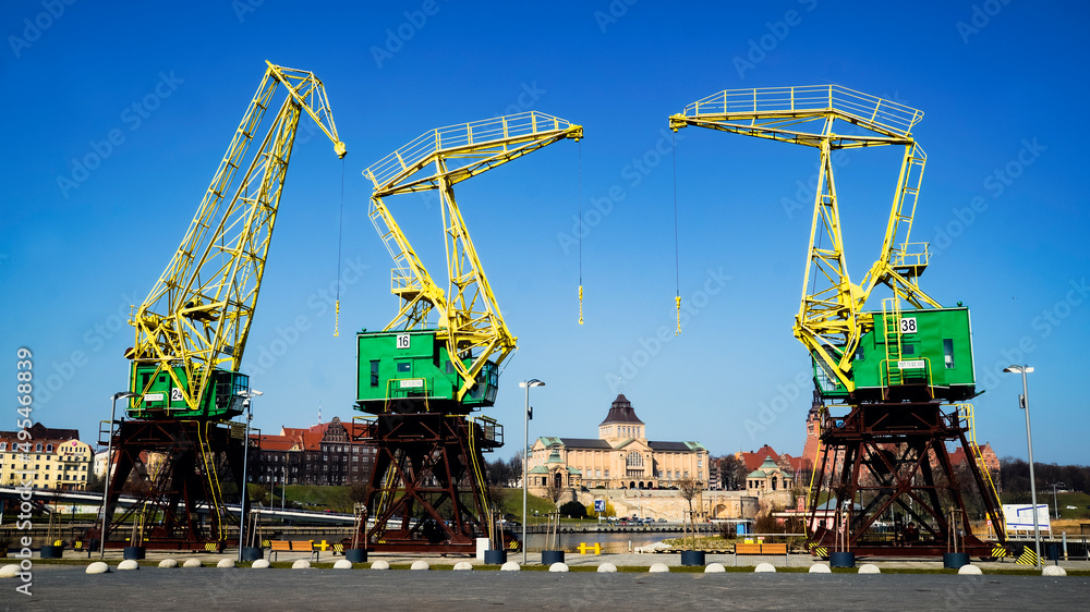 Portowe dźwigi Szczecina na tle intensywnie błękitnego nieba - trzy monumentalne konstrukcje w żółto-zielonej kolorystyce tworzą industrialną orkiestrę, a w tle majestatyczny gmach Wałów Chrobrego przypomina o historycznej potędze miasta