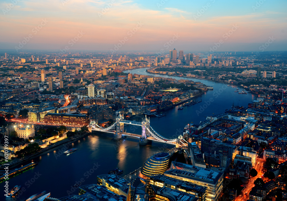 Widok z lotu ptaka na Londyn o zmierzchu, z Tower Bridge na pierwszym planie. Most oświetlony wieczornymi światłami łączy brzegi Tamizy, która wijąc się przez miasto, prowadzi wzrok ku nowoczesnym wieżowcom Canary Wharf w tle. 