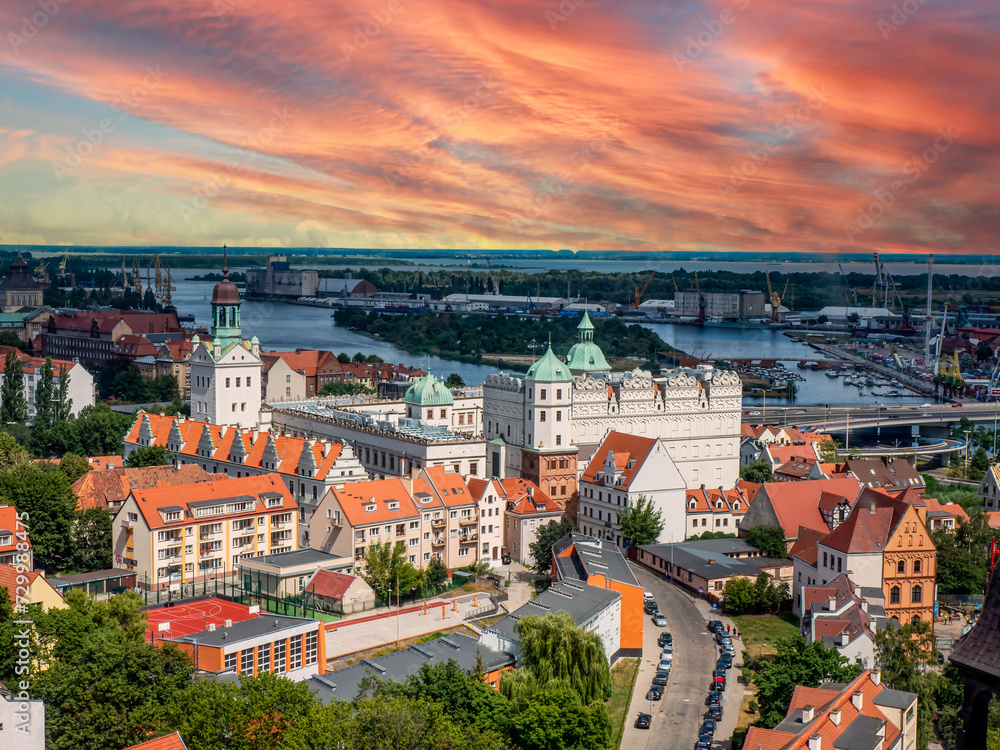 Majestatyczna panorama Szczecina w złotej godzinie - historyczne centrum miasta skąpane w płonącym różowo-pomarańczowym niebie, z charakterystycznymi białymi budynkami i pomarańczowymi dachami, kontrastującymi z industrialnym portem w tle